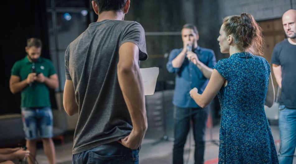 TheaterGruppe von fünf Personen in einem Raum vor der Probe, zwei diskutieren aktiv, andere beobachten oder nutzen Handys.