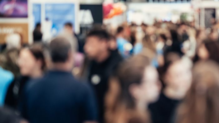 Eine verschwommene Aufnahme von einer Berufsmesse. Viele Menschen gehen einen Gang entlang oder stehen an den Seiten bei den verschiedenen Ständen. Eine verschwommene Aufnahme von einer Berufsmesse. Viele Menschen gehen einen Gang entlang oder stehen an den Seiten bei den verschiedenen Ständen.