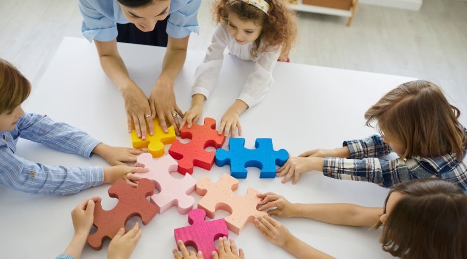 „Mehrere Kinder und eine erwachsene Person sitzen um einen Tisch und setzen gemeinsam große, bunte Puzzleteile zu einem Kreis zusammen.“