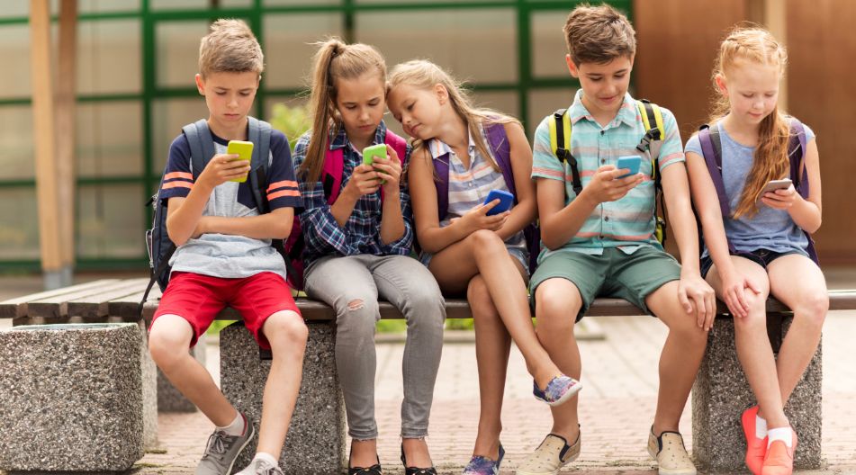 Eine Gruppe von fünf Schulkindern sitzt auf einer Bank im Freien. Alle schauen konzentriert auf ihre Smartphones. Sie tragen Freizeitkleidung und Rucksäcke, was auf eine Schulpause oder den Heimweg hindeutet.