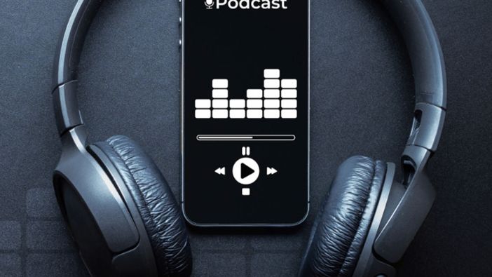 Ein Smartphone liegt mit geöffneter Podcast-App auf einem schwarzem Hintergrund. Um das Smartphone herum sind Kopfhörer. Ein Smartphone liegt mit geöffneter Podcast-App auf einem schwarzem Hintergrund. Um das Smartphone herum sind Kopfhörer.